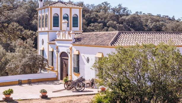 A white building with a bell tower, a tiled roof, and a paved courtyard with a cart and potted plants. - Finca Los Gorriones – Country House with Mountain Views near Seville (El Garrobo)
