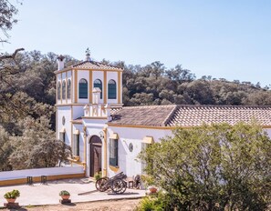 Un bâtiment blanc doté d’un clocher, d’un toit en tuiles et d’une cour pavée où se trouvent une charrette et des plantes en pot.