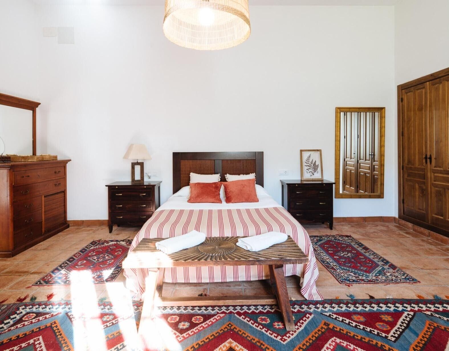 Un dormitorio con una cama, dos mesitas de noche, un aparador de madera, un espejo y alfombras estampadas.