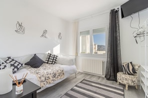 A bedroom with a bed, a small table with a cup and straws, a chair, a window with curtains, and wall art.