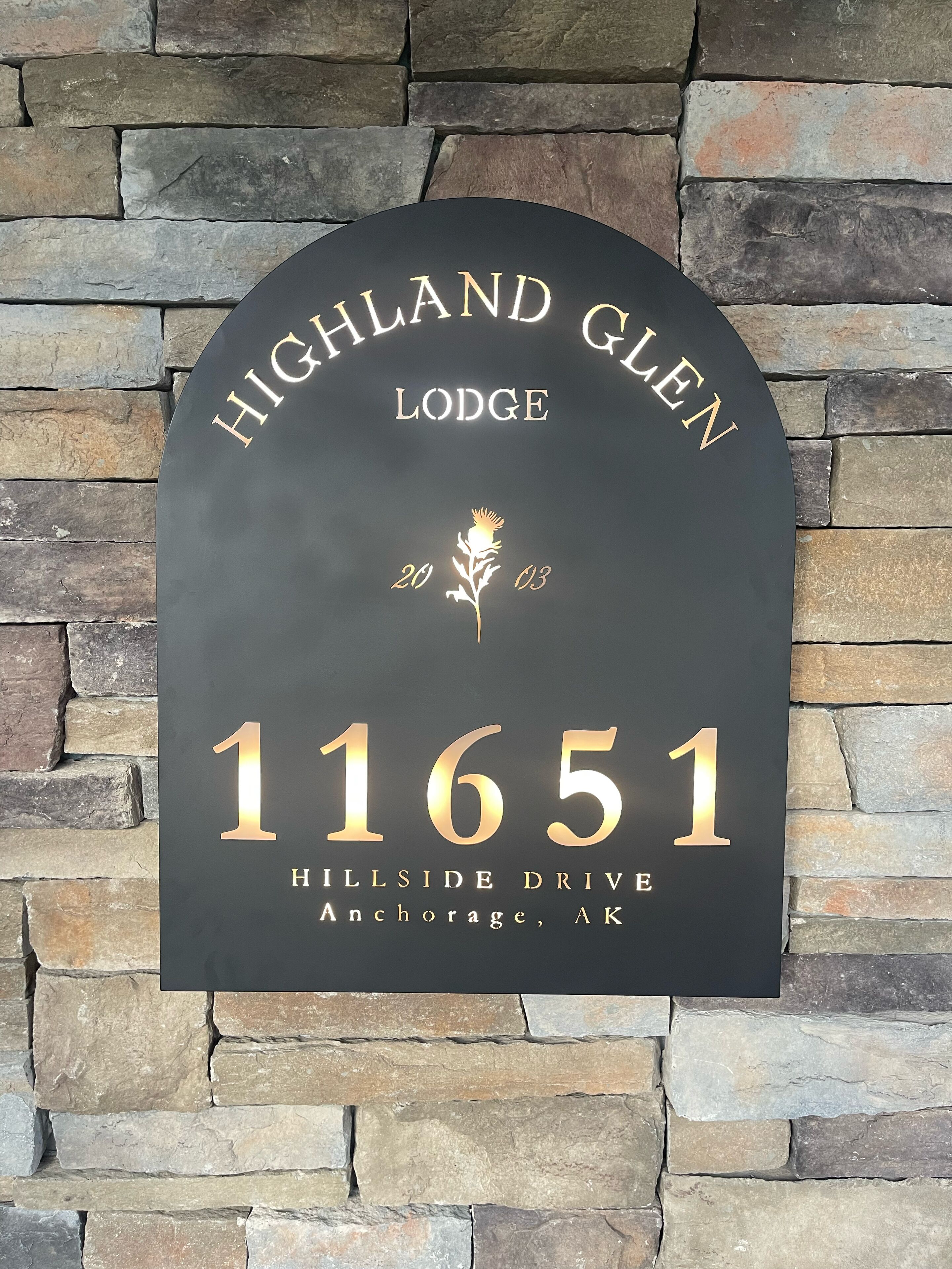 Une plaque noire portant le numéro de la maison et l’inscription « Highland Glen Lodge » ainsi que l’adresse « 11651 Hillside Drive, Anchorage, AK », fixée sur un mur de pierre.