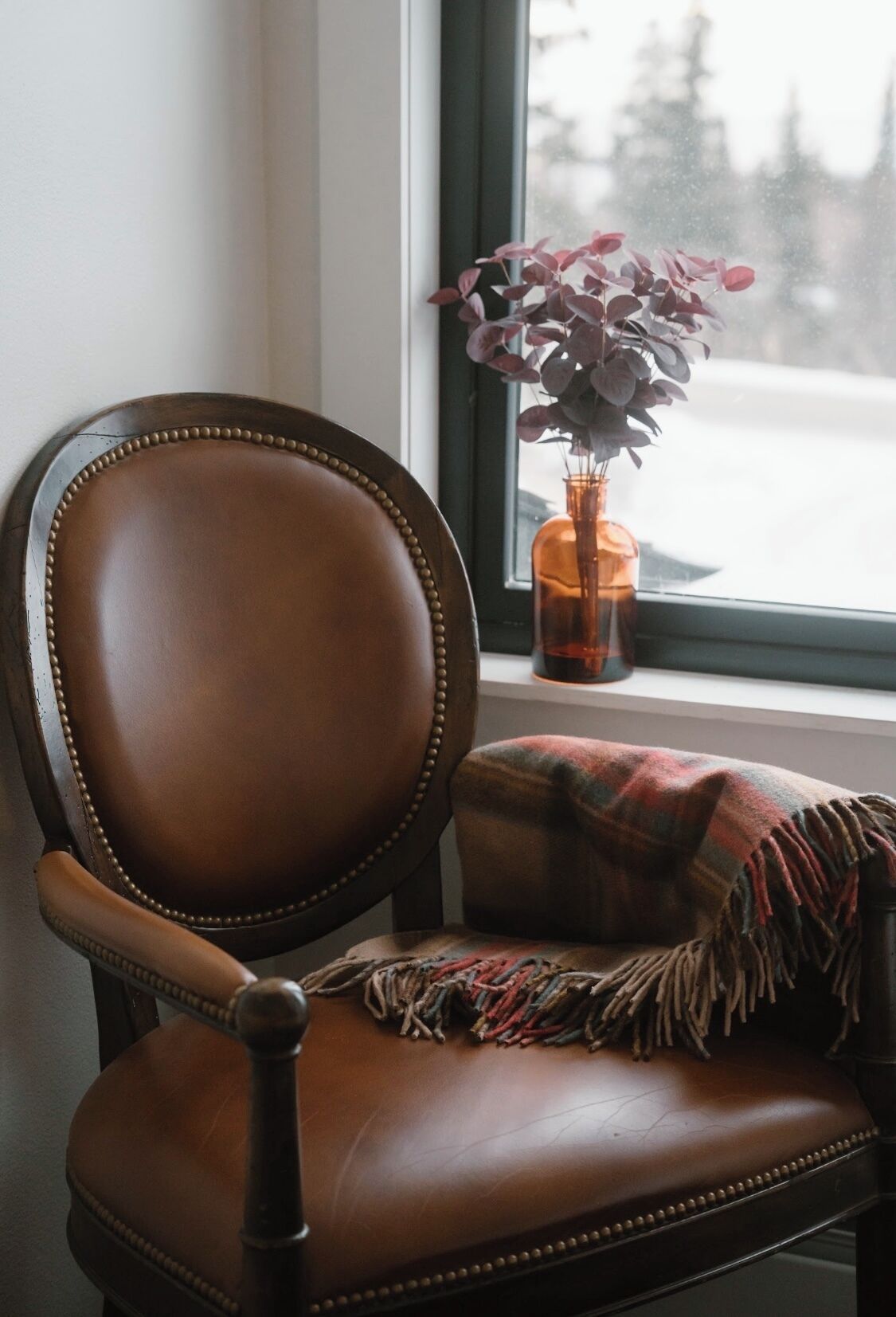 A brown leather chair with a plaid throw blanket, positioned by a window with a view of a snowy landscape.