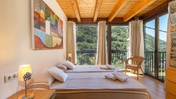 Una stanza con due letti, un soffitto in legno, un'ampia finestra con tende, una poltrona in vimini e un quadro appeso al muro.