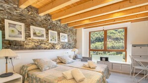 Une chambre avec deux lits, un mur en pierre, des poutres en bois, une grande fenêtre et un bureau.