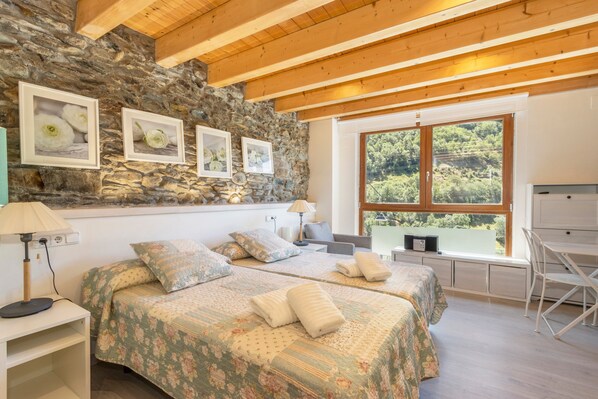 A bedroom with two beds, a stone wall, wooden beams, a large window, and a desk. - Country House 'Casa Guillem 3' with Lake View, Wi-Fi and Air Conditioning (Tavascan)