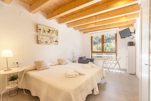 A bedroom with two beds, a desk, a chair, a TV, and a window with a view of greenery.