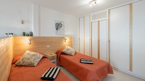 Una habitación con dos camas, cabecero de madera, mesita de noche, lámpara de pared, un cuadro enmarco y un armario.