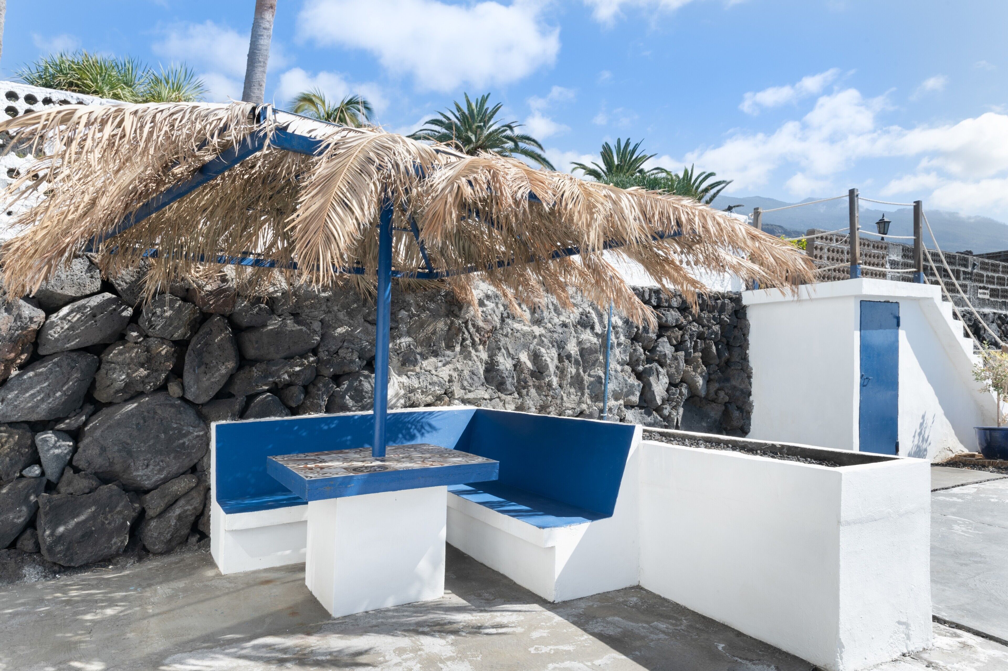 Un espace extérieur moderne, aménagé avec un toit de chaume, des murs en pierre et un banc bleu.