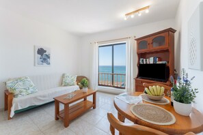 A living room with a sofa, a wooden coffee table, a dining table with chairs, a television, a bookshelf, and a view of the sea through a large window.