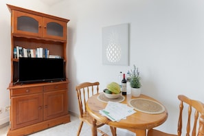 A dining area with a wooden cabinet, a television, a pineapple artwork, a bowl with fruits, a wine bottle, and a potted plant.