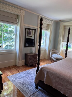 A bedroom with a four-poster bed, a nightstand, a chair, and a large window.