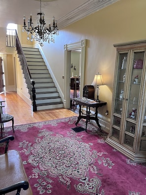 A grand staircase leading to an upper floor, a chandelier, a patterned rug, a table with a lamp, and a glass-fronted cabinet with decorative items. - 1827 Waterfront Mansion, Hot Tub, Fireplace, Huge Deck-River Overlook, fast WiFi (Felts Mills)
