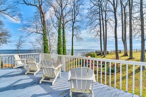 A wooden deck with white Adirondack chairs overlooking a grassy area with trees and a view of the water. - Rising Sun Bay View Cottage W/ Beach Views! (Chesapeake Beach)