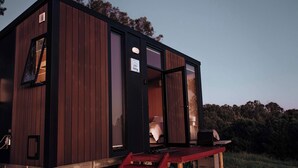 A modern cabin with a wooden exterior, a visible bed inside, and a laptop on a table outside. - Amaroo One by Tiny Away (Minimbah)