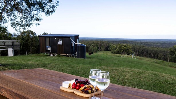 A picnic table with cheese, fruits, and wine glasses, a trailer, and a grassy hill with trees in the background. - Amaroo One by Tiny Away (Minimbah)