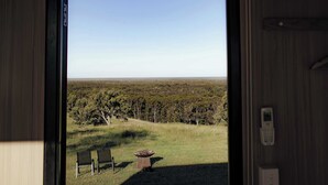 A room with a large window offering a view of a grassy field and trees. There are two chairs and a small table outside the window. - Amaroo One by Tiny Away (Minimbah)