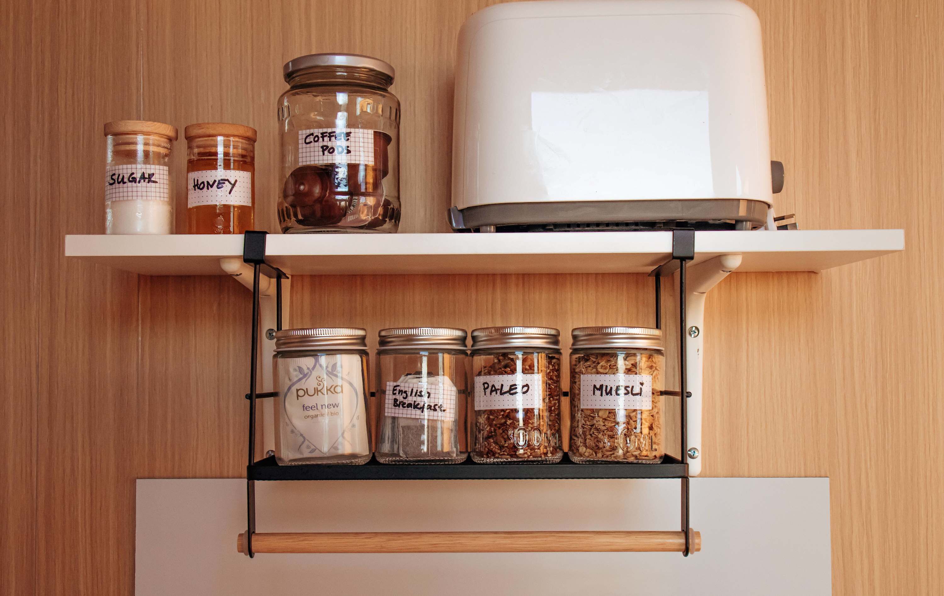 A kitchen with a white appliance, two shelves with labeled jars of sugar, honey, coffee pods, and other ingredients.