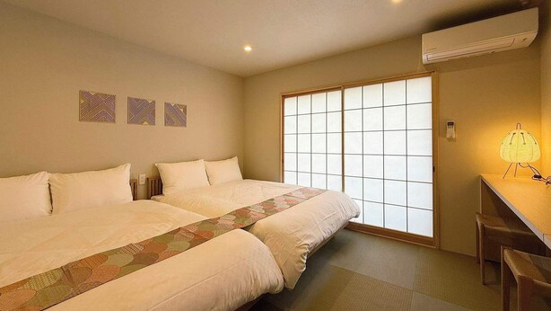 A hotel room with two beds, a desk with a chair, a large window with shoji screens, and a wall-mounted air conditioner. - 2BR, 2 Baths | Vibrant Downtown Near Shopping 21 (Hiroshima)
