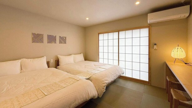 Quarto de hotel com duas camas, um painel deslizante estilo shoji, uma escrivaninha com luminária e um ar condicionado.