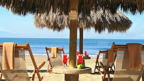 A beach scene with wooden lounge chairs under a thatched umbrella, a wooden post with a sign, and the ocean in the background. - El Tigre: Luxury, Pool, Golf view and near Beaches! (Nuevo Vallarta)
