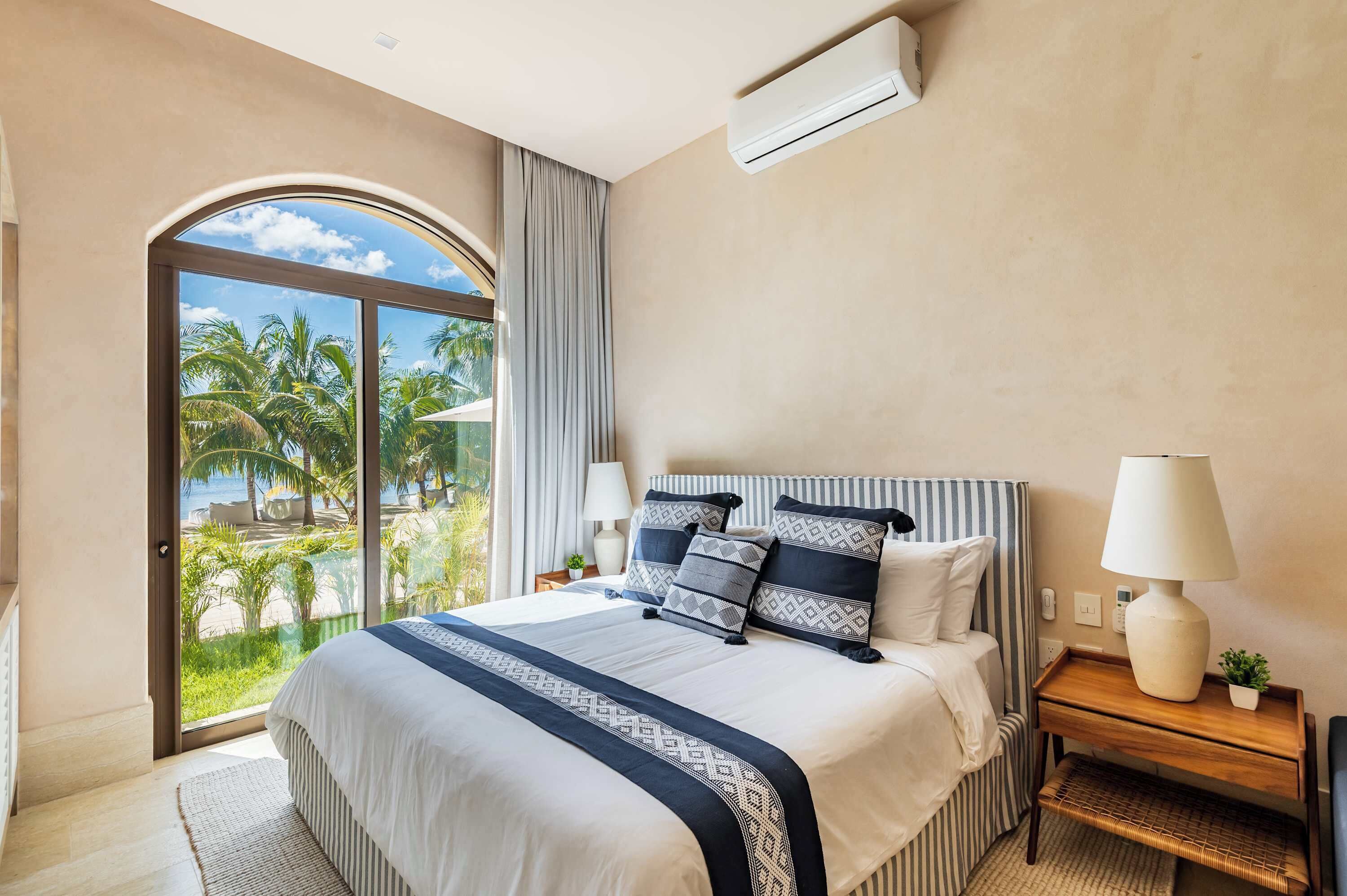 Une chambre d’hôtel avec un grand lit, une table de chevet avec une lampe, et une vue sur des palmiers et une plage, visible à travers une porte vitrée.