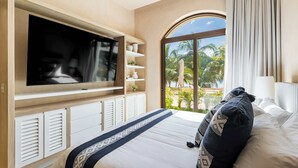 Ein Schlafzimmer mit großem Fernseher, einem Bett mit gemusterten Kissen, einem Fenster mit Blick auf Palmen und einem Balkon.