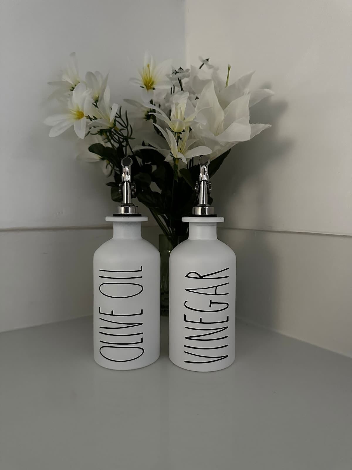 Two white bottles labeled 'Olive Oil' and 'Vinegar' on a white surface with a vase of white flowers in the background.