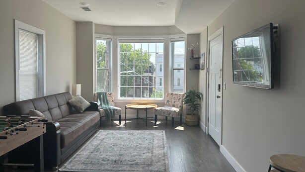 Una sala de estar con un sofá, una mesa de centro y una mesa de futbolín. Hay ventanas amplias con vistas a edificios y árboles.