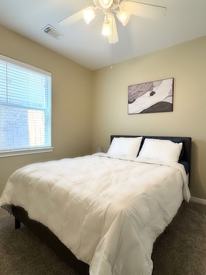 Una camera da letto con un letto, una ventola a soffitto, una finestra con persiane e un quadro astratto appeso al muro.