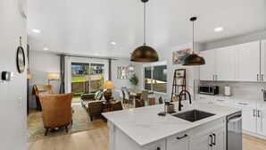 A modern kitchen with white cabinets, a large island with a sink, and pendant lighting. - Gateway to Yellowstone (Livingston)