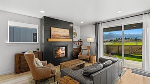 A modern living room with a fireplace, a sofa, a coffee table, and a view of a green landscape through large windows. - Gateway to Yellowstone (Livingston)