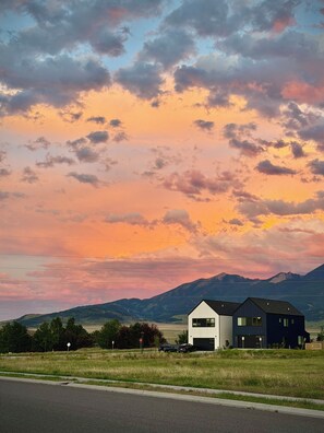 A modern two-story house with a flat roof and large windows, situated on a grassy field with a mountain range in the background. - Gateway to Yellowstone (Livingston)