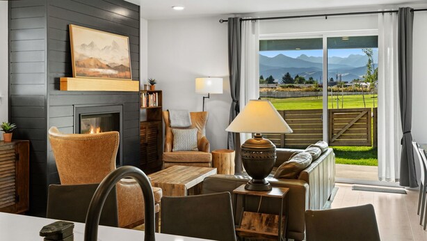 A modern living room with a fireplace, comfortable seating, and a view of a green landscape through large windows. - Gateway to Yellowstone (Livingston)