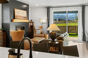 A modern living room with a fireplace, comfortable seating, and a view of a green landscape through large windows. - Gateway to Yellowstone (Livingston)