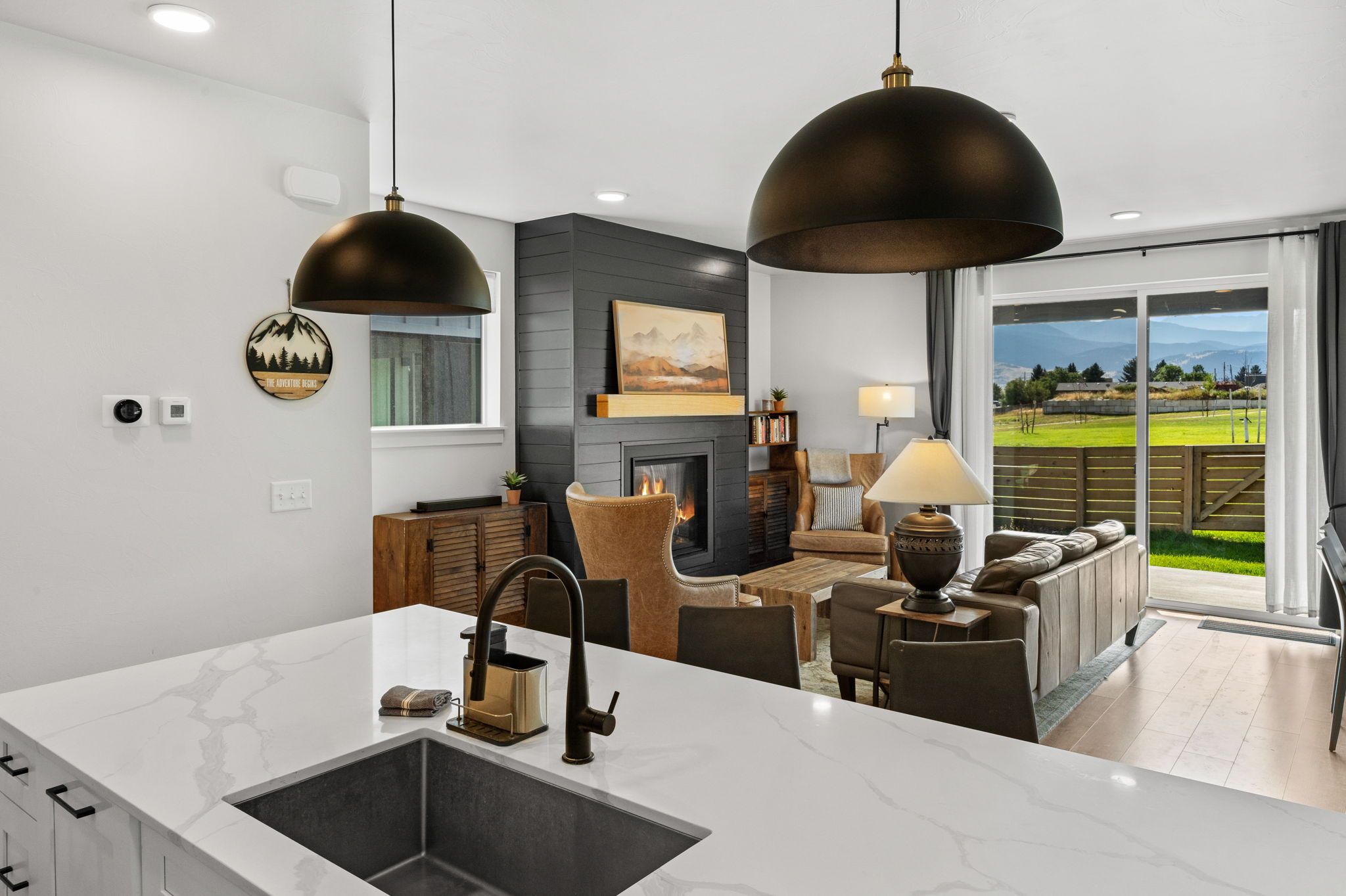 A modern kitchen with a marble countertop, black faucet, and pendant lights. There is a fireplace with a painting above it, and a seating area with a sofa and coffee table.