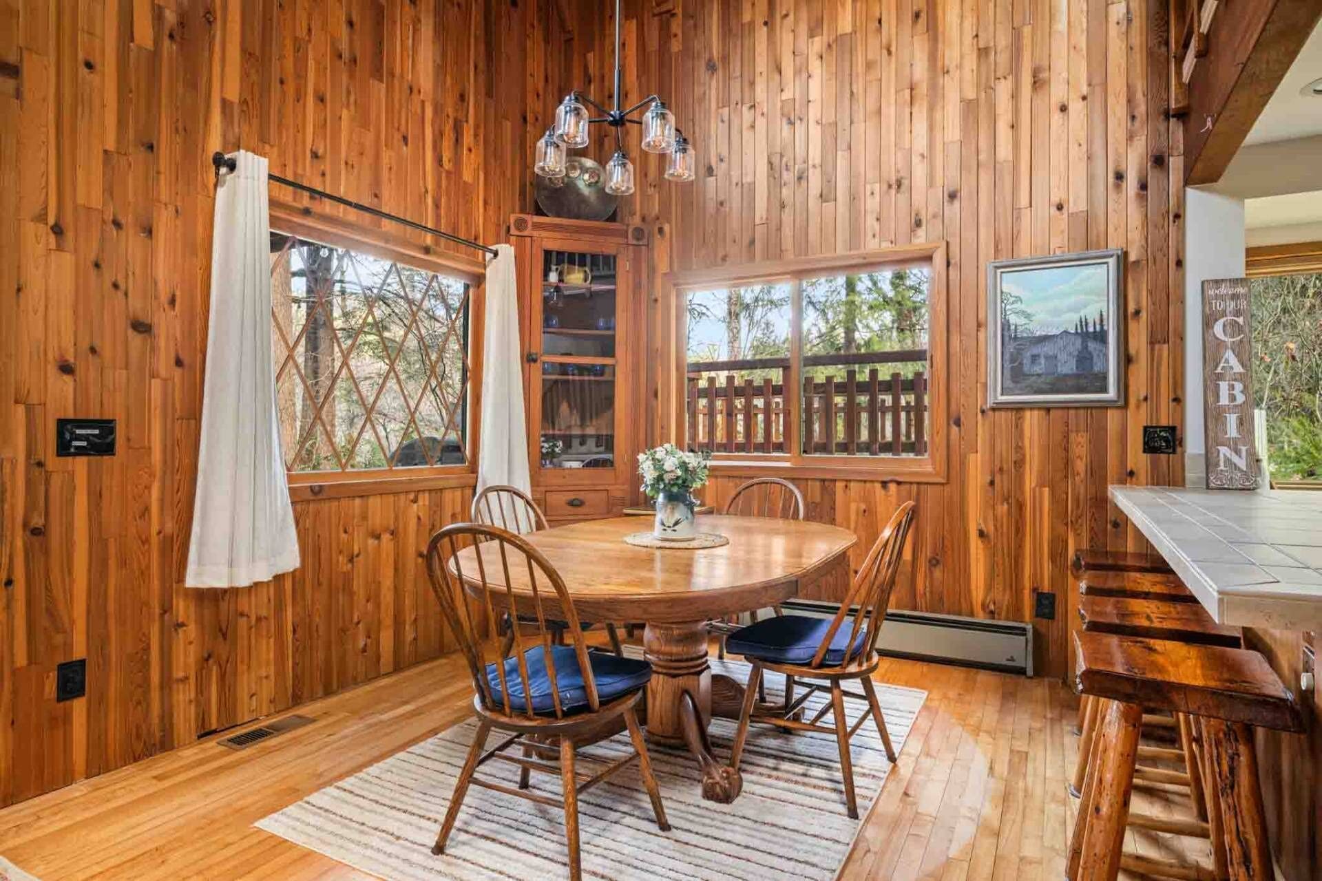 Ein Holzblock-Interieur mit einem runden Esstisch, vier Stühlen mit blauen Kissen, einer Kristallleuchtre und einem großen Fenster mit Blick auf Bäume.
