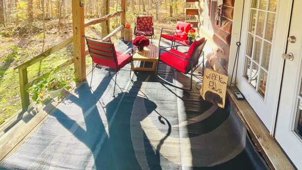 Een houten terras met een overdekte zithoek, een bord met de tekst 'Welcome to the Cabin', en een uitzicht op het omliggende bos.