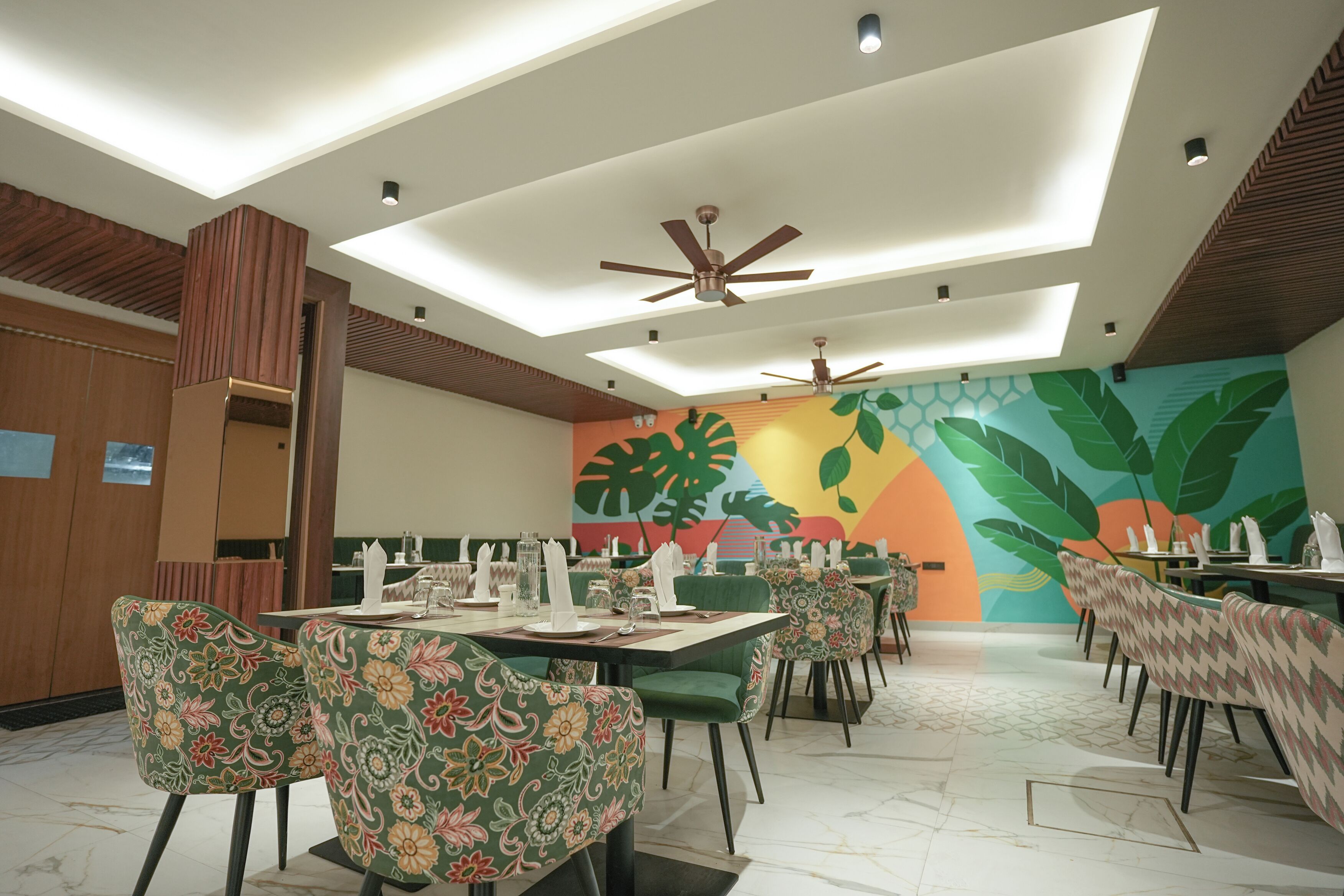 Un restaurante con sillas tapizadas en verde, ventiladores de techo y un mural colorido en la pared.