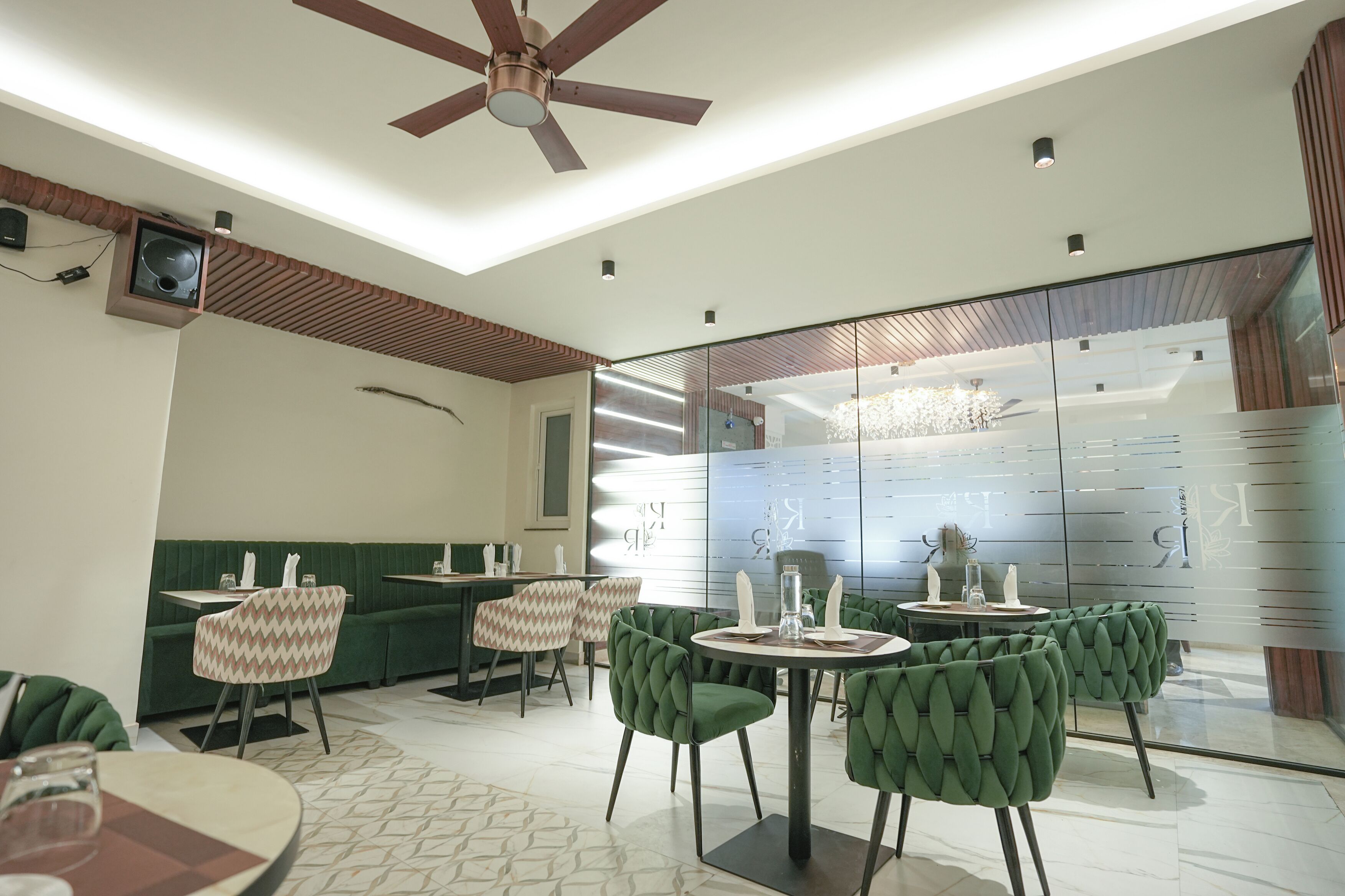 A modern restaurant with green upholstered chairs, a ceiling fan, and a glass partition wall.