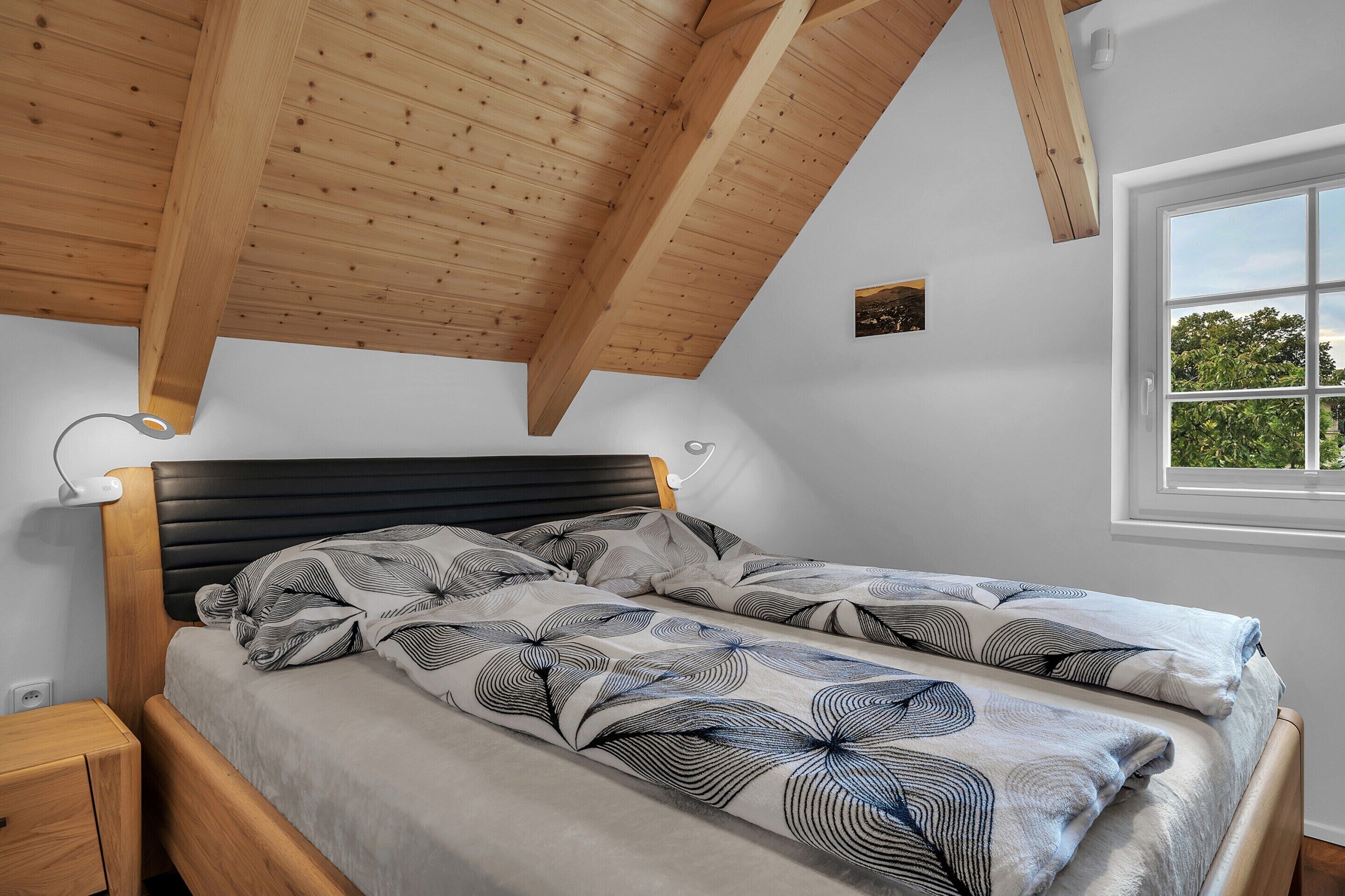 A bedroom with a wooden ceiling, a bed with patterned bedding, a bedside table, and a window with a view of greenery.