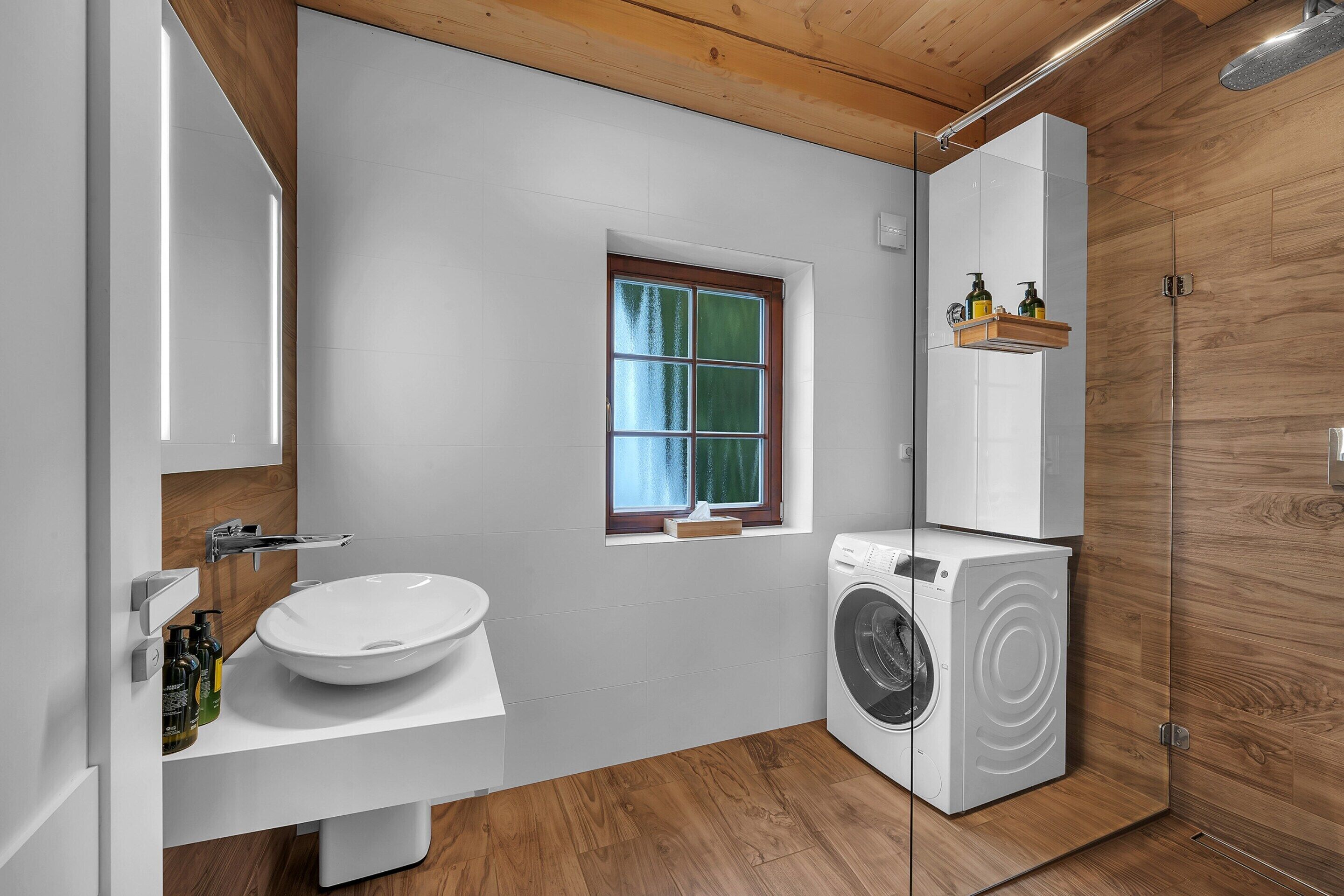 A modern bathroom with a wooden floor, a white sink, a built-in washing machine, and a window with a view.