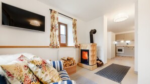 A cozy living room with a fireplace, a sofa with floral cushions, a television mounted on the wall, and a kitchen area visible through an open door. - Villa in Varvazov With Garden and Barbecue (Varvažov)