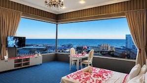 A hotel room with a large bay window, a bed with rose petals, a chandelier, a TV, and a desk with a chair. - Olive Garden Hotel Baku (Baku)