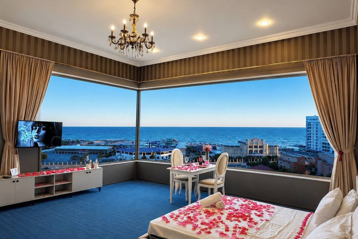 A hotel room with a large bay window, a bed with rose petals, a chandelier, a TV, and a desk with a chair.