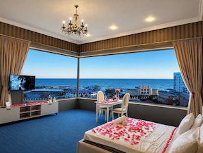Een hotelkamer met een groot erkerraam, een bed met rozenblaadjes, een kroonluchter, een televisie en een bureau met een stoel.