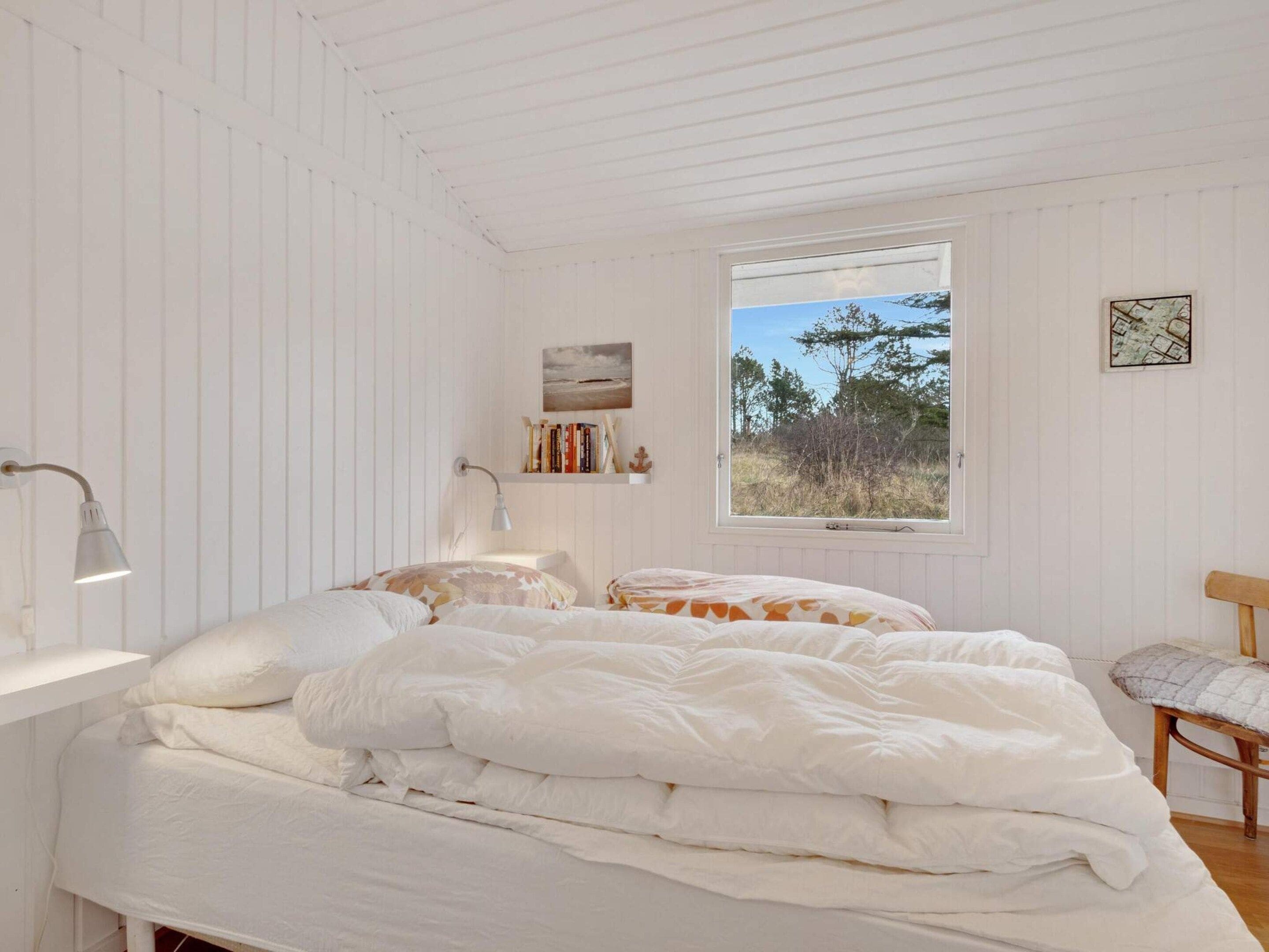 A bedroom with a bed, a desk, a chair, a window with a view of trees, and a shelf with books and a picture.