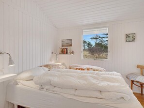 A bedroom with a bed, a desk, a chair, a window with a view of trees, and a shelf with books and a picture. - 5 Star Holiday Home in Lokken (Lokken)