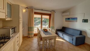 A compact living space with a kitchenette, a dining area with a table and chairs, a sofa, and a balcony with a view of greenery. - Ancora Sardinia Seaside Apartment (Badesi)