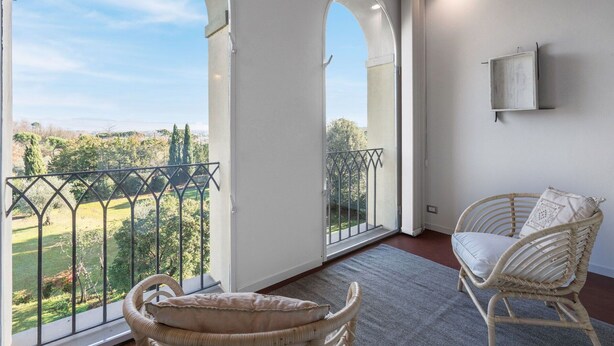 Un balcon donnant sur un paysage verdoyant, une chaise en osier et un mur blanc orné d’une armoire fixée au mur.