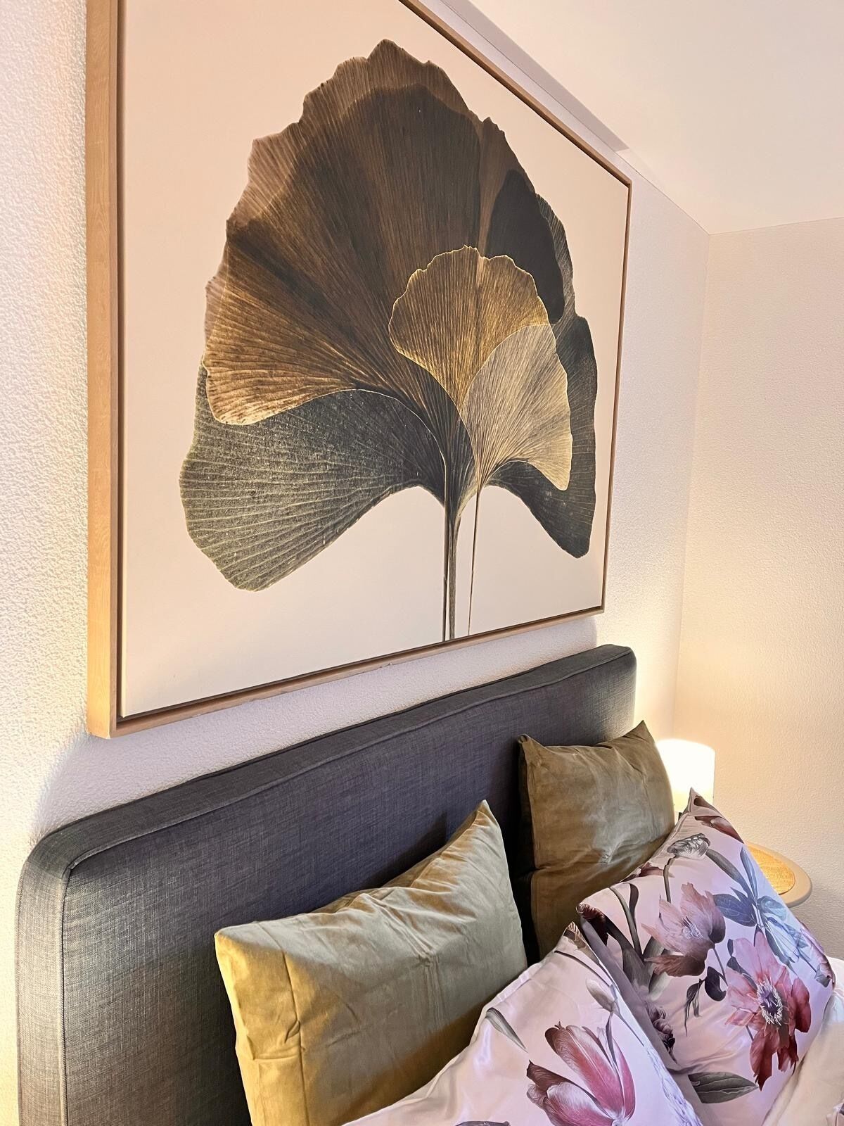 Een lijst met een bloem aan een muur boven een bed met een grijs hoofdeinde en kussens met bloemenprint.